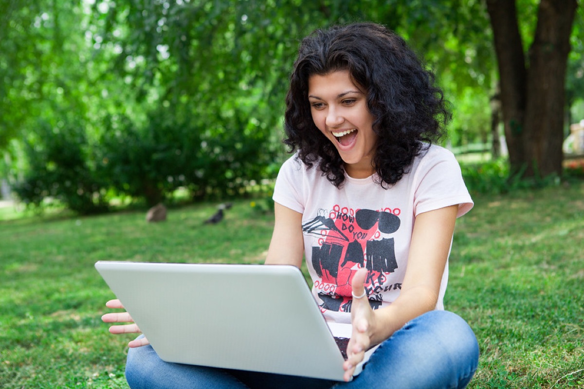 A student laughing at her laptop, sitting on the grass